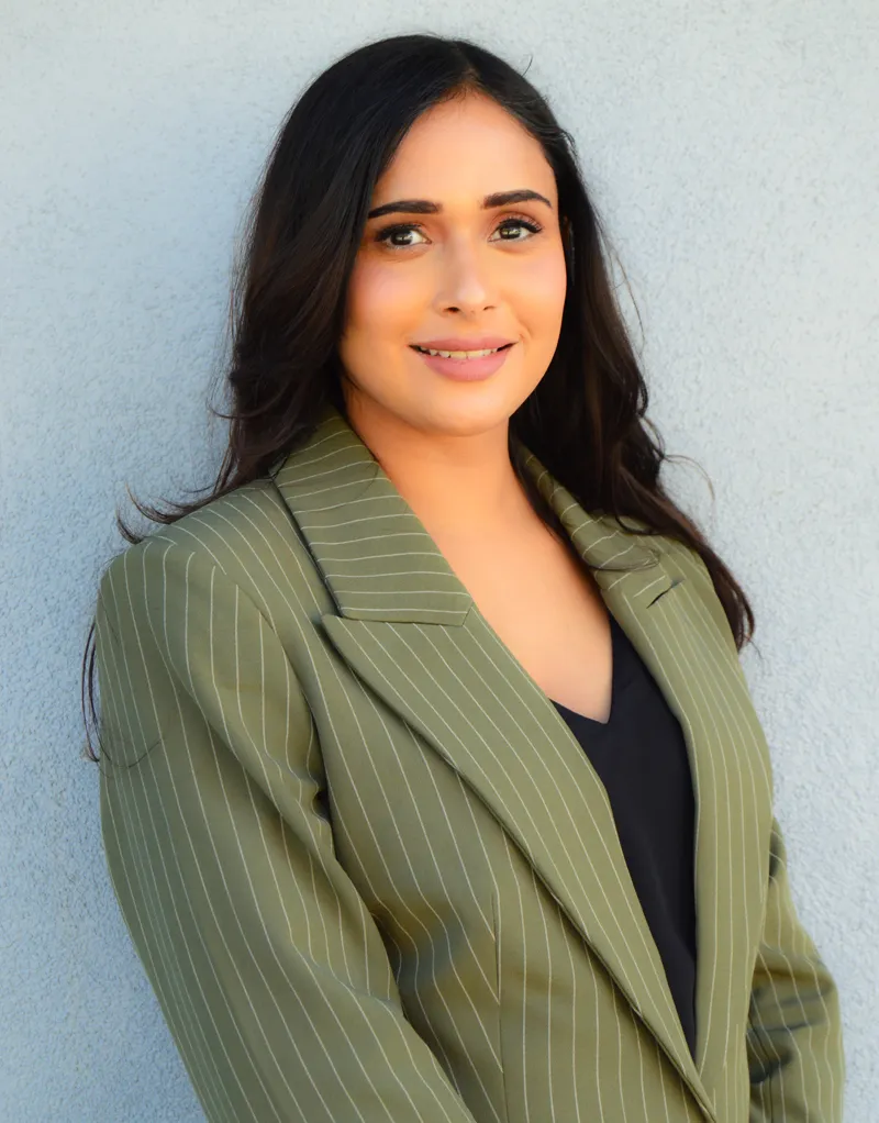 Dr. Gurpreet Kaur, female dentist at Fairfield Dentistry & Implants, wearing green pinstripe blazer, smiling in front of light blue wall